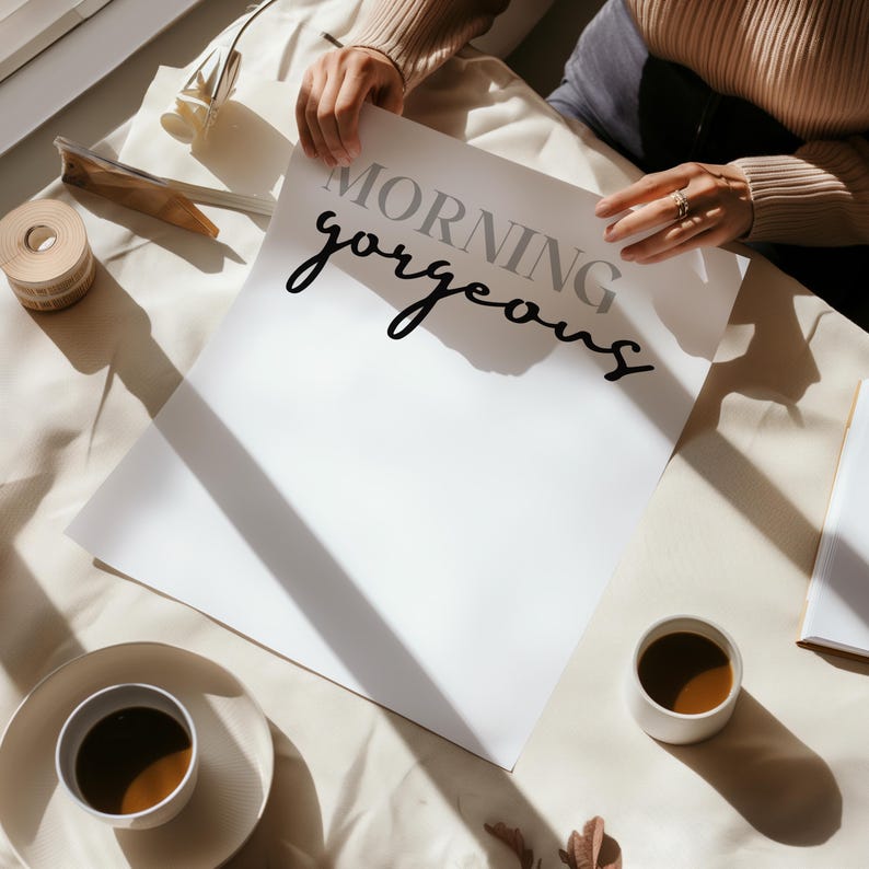 May include: A white card with the text "Morning gorgeous" written in black cursive script. The card is lying on a white surface with a cup of coffee and a shadow.