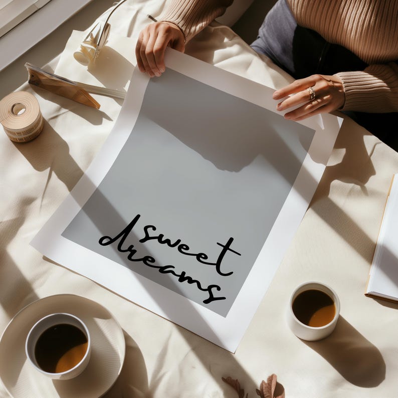 May include: A white poster with the text "Sweet Dreams" written in black cursive script. The poster is lying on a white surface with a cup of coffee and a shadow.