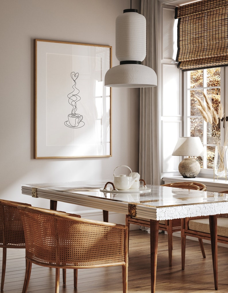 May include: A white ceramic pendant light hangs over a dining table with a white marble top and gold accents. Two wicker chairs are placed on either side of the table. A framed print with a line drawing of a coffee cup with hearts for steam hangs on the wall behind the table.