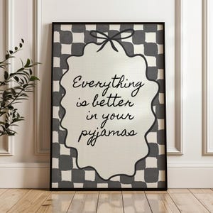 Może przedstawiać: Czarna oprawiona grafika ścienna z napisem "Everything is better in your pyjamas". Grafika przedstawia kremowy kształt z falistymi krawędziami z czarnym napisem na czarno-białym tle w szachownicę.