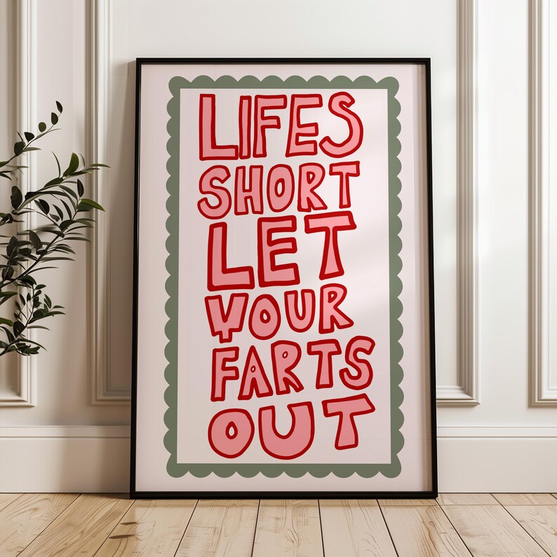 May include: A framed art print with the text "LIFES SHORT LET YOUR FARTS OUT" in red, outlined in a darker red. The text is on a light pink background, framed by a green scalloped border and a black frame. The print is displayed on a wooden floor.