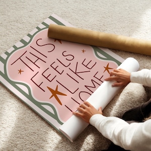 Puede incluir: P&oacute;ster enrollado con el texto "THIS FEELS LIKE HOME" en una fuente caprichosa, con un dise&ntilde;o rosa y verde y detalles de estrellas. Un segundo p&oacute;ster enrollado est&aacute; al lado.