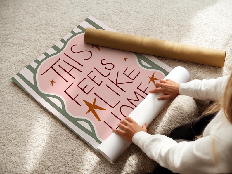 Puede incluir: P&oacute;ster enrollado con el texto "THIS FEELS LIKE HOME" en una fuente caprichosa, con un dise&ntilde;o rosa y verde y detalles de estrellas. Un segundo p&oacute;ster enrollado est&aacute; al lado.