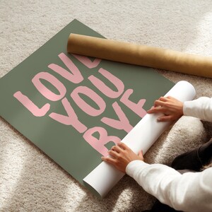 May include: An olive green poster with the words "LOVE YOU BYE" in pink lettering. A rolled-up poster and a brown cardboard tube are also visible. The poster is on a light-coloured carpet.