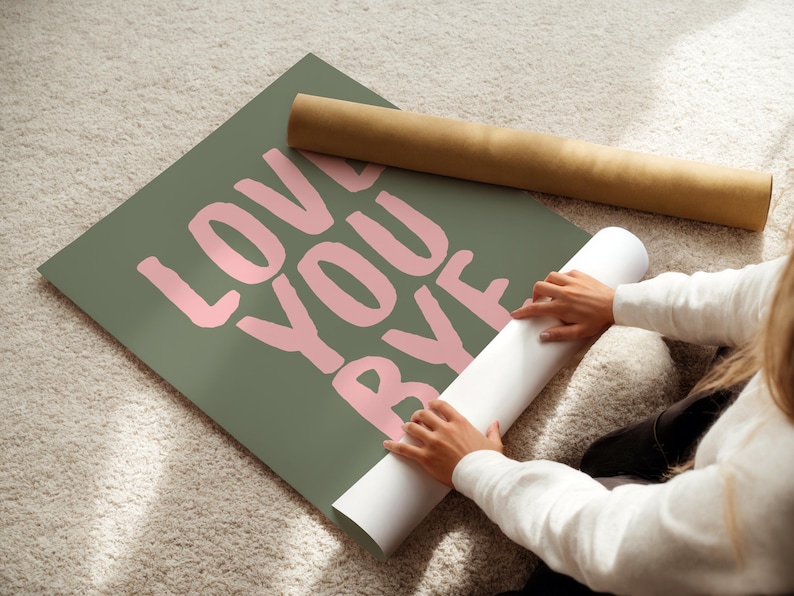 May include: An olive green poster with the words "LOVE YOU BYE" in pink lettering. A rolled-up poster and a brown cardboard tube are also visible. The poster is on a light-coloured carpet.