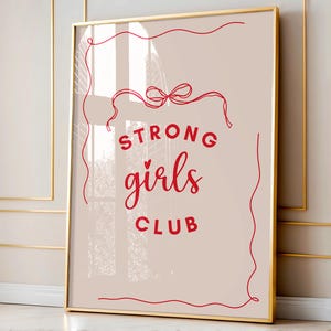 Op de afbeelding: Een gouden ingelijste print met een roze achtergrond en rode tekst die "STRONG girls CLUB" luidt met een rood lint en strik bovenaan.