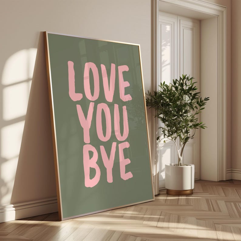 May include: A framed print featuring the words "LOVE YOU BYE" in pink on a green background. The artwork is propped against a wall in a room, with a plant in a white pot nearby.