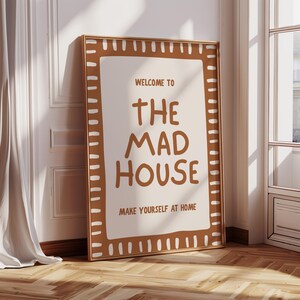 May include: A framed poster with the text "WELCOME TO THE MAD HOUSE MAKE YOURSELF AT HOME". The poster has a brown border with white accents and is leaning against a wall. The background includes a window and a curtain.