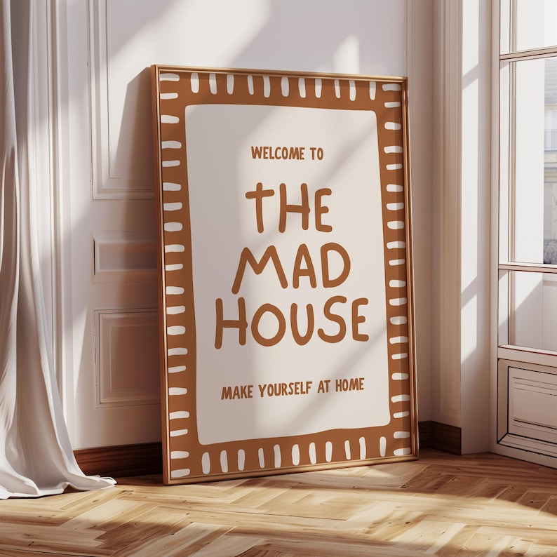 May include: A framed poster with the text "WELCOME TO THE MAD HOUSE MAKE YOURSELF AT HOME". The poster has a brown border with white accents and is leaning against a wall. The background includes a window and a curtain.