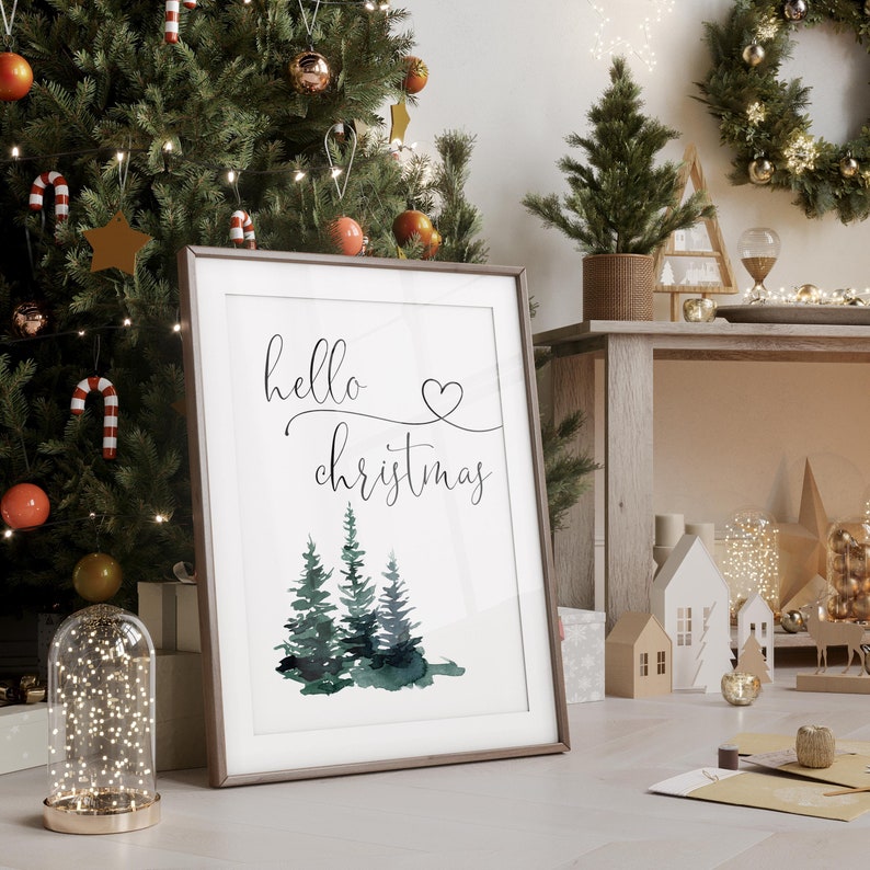 May include: A framed print with the text "hello christmas" in a cursive font with a small heart above the word "christmas". The print features a watercolor illustration of three green pine trees.
