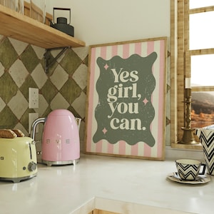 May include: A kitchen scene featuring a pink electric kettle and a yellow toaster. A framed print with the text "Yes girl, you can" is displayed. A cup and saucer with a black and white geometric design are also visible.