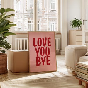 May include: A framed pink art print with the words "LOVE YOU BYE" in bold red letters. The artwork is displayed in a bright room with a large window, a plant, and a cardboard box.