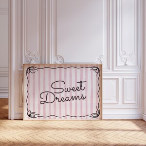 Op de afbeelding: Een ingelijste print met de woorden "Sweet Dreams" in elegant schrift. De print heeft een roze en witte gestreepte achtergrond met een decoratieve zwarte rand. De lijst is licht van kleur, en de print wordt getoond in een kamer met witte muren en een houten vloer.