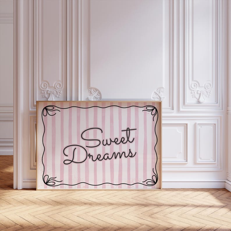 Op de afbeelding: Een ingelijste print met de woorden "Sweet Dreams" in elegant schrift. De print heeft een roze en witte gestreepte achtergrond met een decoratieve zwarte rand. De lijst is licht van kleur, en de print wordt getoond in een kamer met witte muren en een houten vloer.
