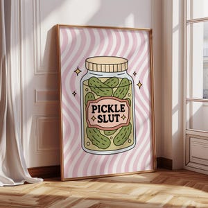 May include: Framed art print with a cartoon jar of pickles. The jar has a beige lid and a label reading "PICKLE SLUT". The background features pink and white stripes. The illustration has a retro style.