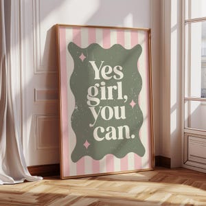 May include: A framed print with the text "Yes girl, you can." in a distressed font. The text is on a sage green, wavy-edged shape, set against a pink and white striped background. The frame is gold.