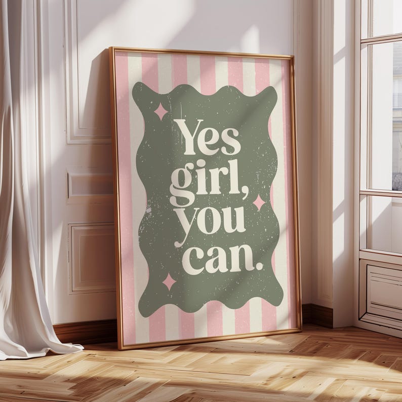 May include: A framed print with the text "Yes girl, you can." in a distressed font. The text is on a sage green, wavy-edged shape, set against a pink and white striped background. The frame is gold.