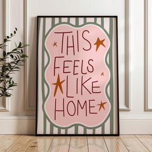 Puede incluir: Impresi&oacute;n art&iacute;stica enmarcada con la frase "THIS FEELS LIKE HOME" en un estilo manuscrito. El texto es rojo oscuro sobre una forma rosa con bordes ondulados, con estrellas doradas. El fondo tiene rayas verticales verdes y blancas.