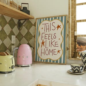 Puede incluir: Escena de cocina con una tostadora amarilla clara, una tetera el&eacute;ctrica rosa y un letrero enmarcado que dice "THIS FEELS LIKE HOME". Tambi&eacute;n se ve una taza y un platillo con estampado en blanco y negro.