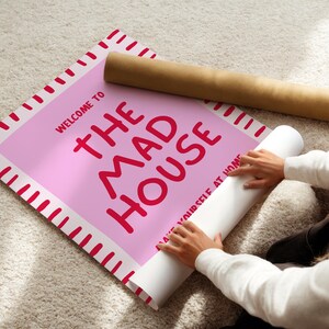 May include: A pink poster with the words "WELCOME TO THE MAD HOUSE" in red lettering. The poster is being unrolled, revealing the text "MAKE YOURSELF AT HOME". A brown cardboard tube is next to the poster.