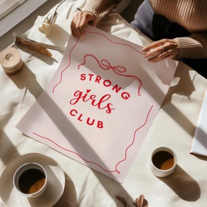 Op de afbeelding: Een roze poster met rode tekst die "STRONG girls CLUB" luidt met een rood touwontwerp rond de tekst.