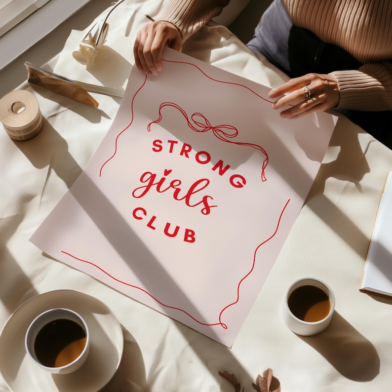 Op de afbeelding: Een roze poster met rode tekst die "STRONG girls CLUB" luidt met een rood touwontwerp rond de tekst.
