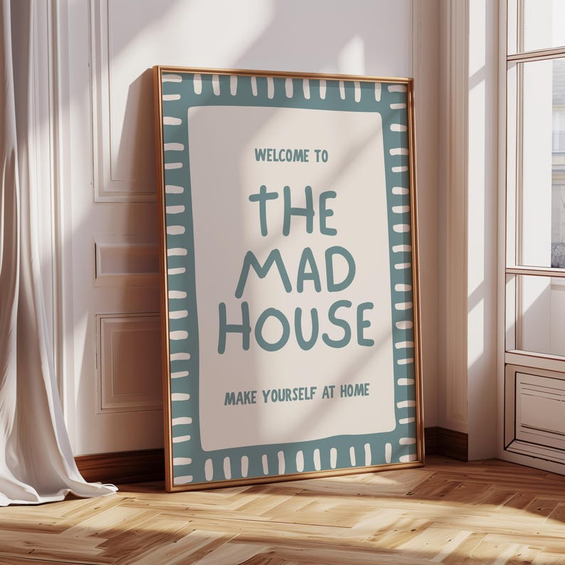 May include: Framed poster with the text "WELCOME TO THE MAD HOUSE MAKE YOURSELF AT HOME" in teal lettering. The poster has a teal border with white dashes and a gold frame. It leans against a white wall and wooden floor.