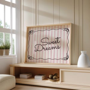 Op de afbeelding: Een ingelijste print met de woorden "Sweet Dreams" in elegant schrift. De achtergrond heeft roze en witte verticale strepen, omlijst door een decoratieve zwarte rand. Het frame heeft een lichte houtkleur.