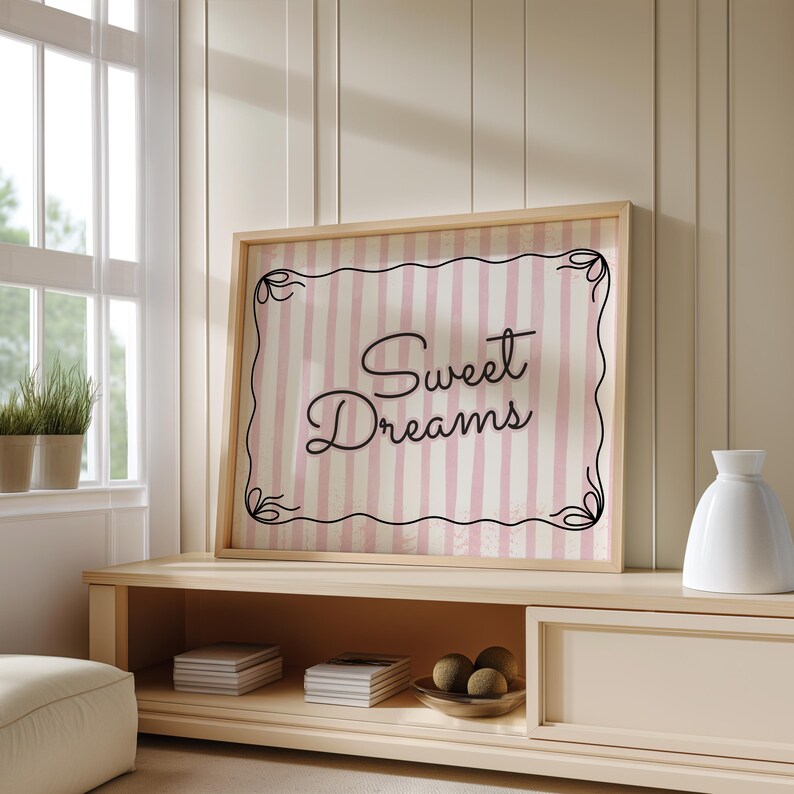 Op de afbeelding: Een ingelijste print met de woorden "Sweet Dreams" in elegant schrift. De achtergrond heeft roze en witte verticale strepen, omlijst door een decoratieve zwarte rand. Het frame heeft een lichte houtkleur.