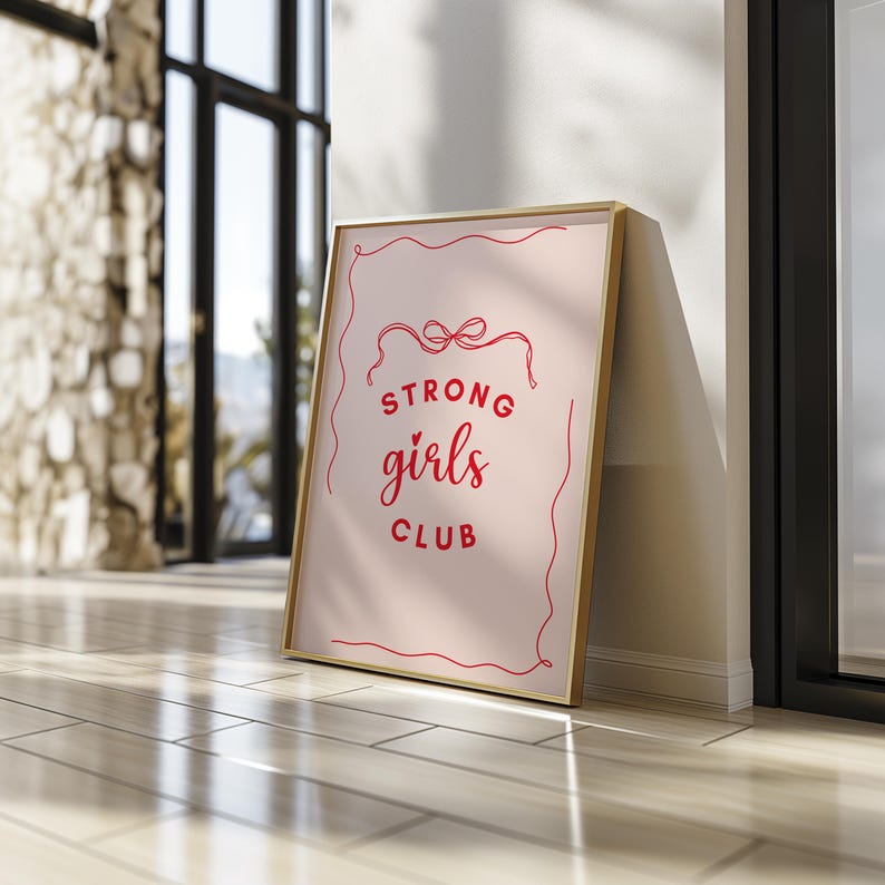 Op de afbeelding: Een gouden ingelijste print met een roze achtergrond en rode tekst die "Strong girls club" luidt.