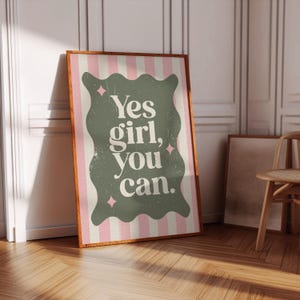 May include: A framed poster with the text "Yes girl, you can." The poster has a green, wavy-edged shape with white text on a pink and white striped background. The frame is brown.