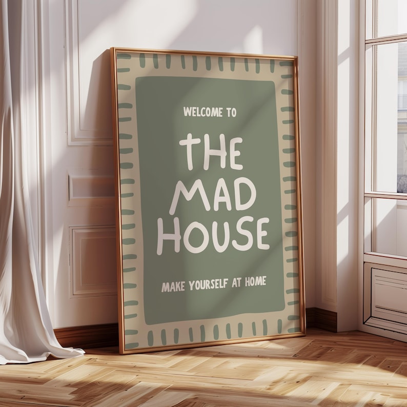 May include: A framed poster with the text "WELCOME TO THE MAD HOUSE MAKE YOURSELF AT HOME" in white on a green background. The poster has a tan border and a gold frame, leaning against a white wall and wooden floor.