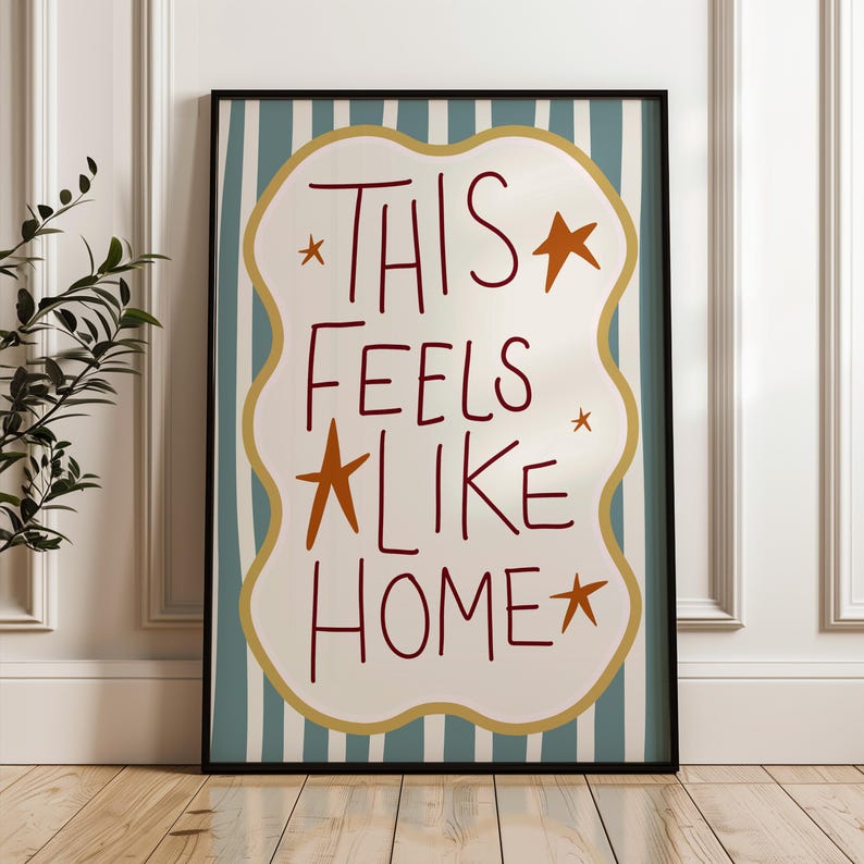 Puede incluir: Un cuadro enmarcado con la frase "THIS FEELS LIKE HOME" en un estilo manuscrito. El texto es de color rojo oscuro, con estrellas naranjas. El fondo tiene una forma ondulada blanca con un borde dorado, sobre un fondo a rayas azules y blancas.