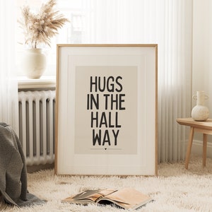 May include: A framed print with the words "HUGS IN THE HALL WAY" in black text. The print is in a light-colored frame and is displayed in a room with a fluffy rug, a vase with dried flowers, and a small wooden table.
