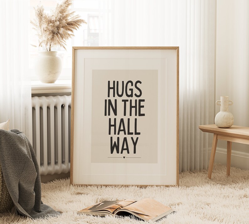 May include: A framed print with the words "HUGS IN THE HALL WAY" in black text. The print is in a light-colored frame and is displayed in a room with a fluffy rug, a vase with dried flowers, and a small wooden table.