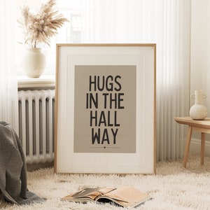 May include: A framed print with the words "HUGS IN THE HALL WAY" in black text on a tan background. The print is in a room with a fluffy white rug, a vase with dried flowers, and a wooden bench.