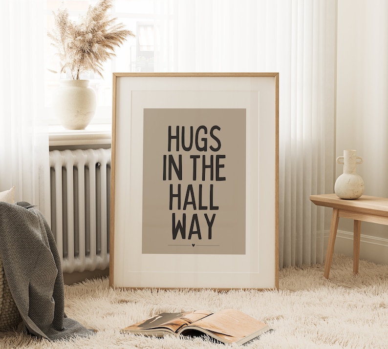 May include: A framed print with the words "HUGS IN THE HALL WAY" in black text on a tan background. The print is in a room with a fluffy white rug, a vase with dried flowers, and a wooden bench.