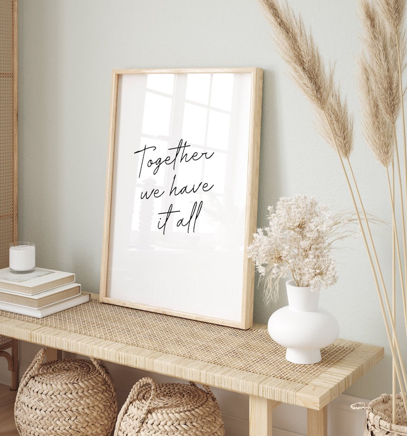 May include: A framed print with the text "Together we have it all" in black script. The print is displayed on a light wood bench with woven detailing, alongside a white vase with dried flowers and decorative baskets.