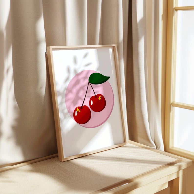 Op de afbeelding: Een ingelijst print met een roze cirkel en twee rode kersen met groene stelen en bladeren. De kersen hebben een glanzende, glinsterende textuur.