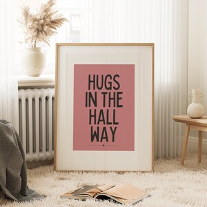 May include: A framed art print with the words "HUGS IN THE HALL WAY" in black on a dusty rose background. The print is in a light wood frame and is displayed in a cozy room with a fluffy rug and decorative vase.