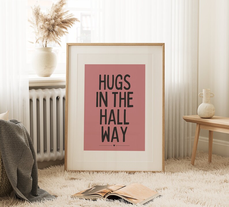 May include: A framed art print with the words "HUGS IN THE HALL WAY" in black on a dusty rose background. The print is in a light wood frame and is displayed in a cozy room with a fluffy rug and decorative vase.