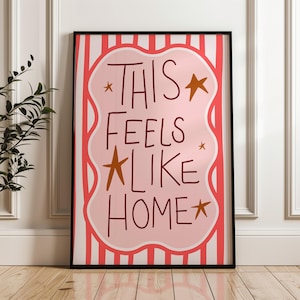 Puede incluir: Una impresi&oacute;n art&iacute;stica enmarcada con las palabras "THIS FEELS LIKE HOME" en un estilo manuscrito. El fondo tiene un patr&oacute;n de rayas rosas y blancas con una forma de nube rosa en el centro. Estrellas doradas est&aacute;n dispersas alrededor del texto.