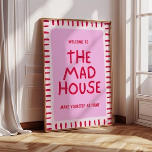 May include: A framed pink poster with the words "WELCOME TO THE MAD HOUSE MAKE YOURSELF AT HOME" in red lettering. The poster has a gold frame and is leaning against a white wall and wooden floor.