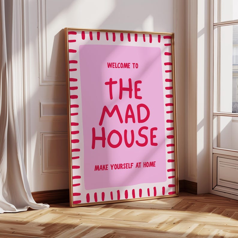 May include: A framed pink poster with the words "WELCOME TO THE MAD HOUSE MAKE YOURSELF AT HOME" in red lettering. The poster has a gold frame and is leaning against a white wall and wooden floor.