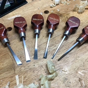 May include: Six wood carving tools with polished dark brown wooden palm handles and silver metal ferrules. Each handle is stamped with the text RAMELSON USA. The tools feature various steel blade shapes, including straight, angled, and curved tips.