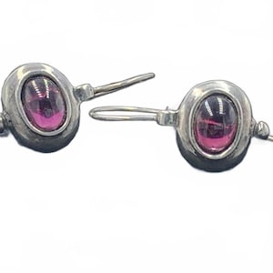 May include: A pair of silver earrings with oval, magenta-colored gemstones. The earrings have a hook closure and a small, decorative ball at the end of the post. The gemstones are set in a bezel setting, and the metal has a slightly oxidized appearance.