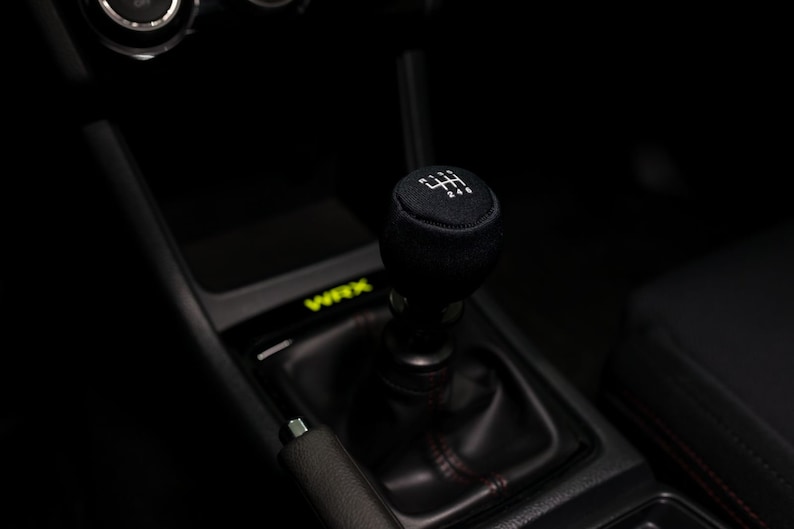 Op de afbeelding: Close-up van een zwarte leren handgeschakelde versnellingspookknop met een wit versnellingschema. De knop bevindt zich in een zwart auto-interieur.