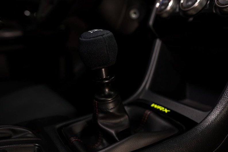 Op de afbeelding: Een zwarte leren versnellingspookknop met een zwarte stoffen hoes. De versnellingspook bevindt zich in het interieur van een auto.