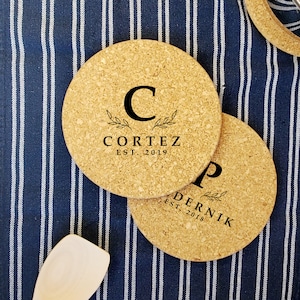 May include: Two round cork coasters with black text and leaf accents. One coaster features the letter "C" and the name "CORTEZ EST. 2019". The other coaster has the letter "P" and the name "DERNIk EST. 2018". The coasters are on a blue and white striped surface.