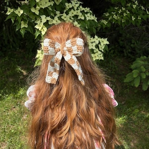 May include: A cream and light brown checkered crochet hair bow, set against long, wavy auburn hair. The bow is the focal point, adding a touch of whimsy. The background features green foliage and a floral dress.